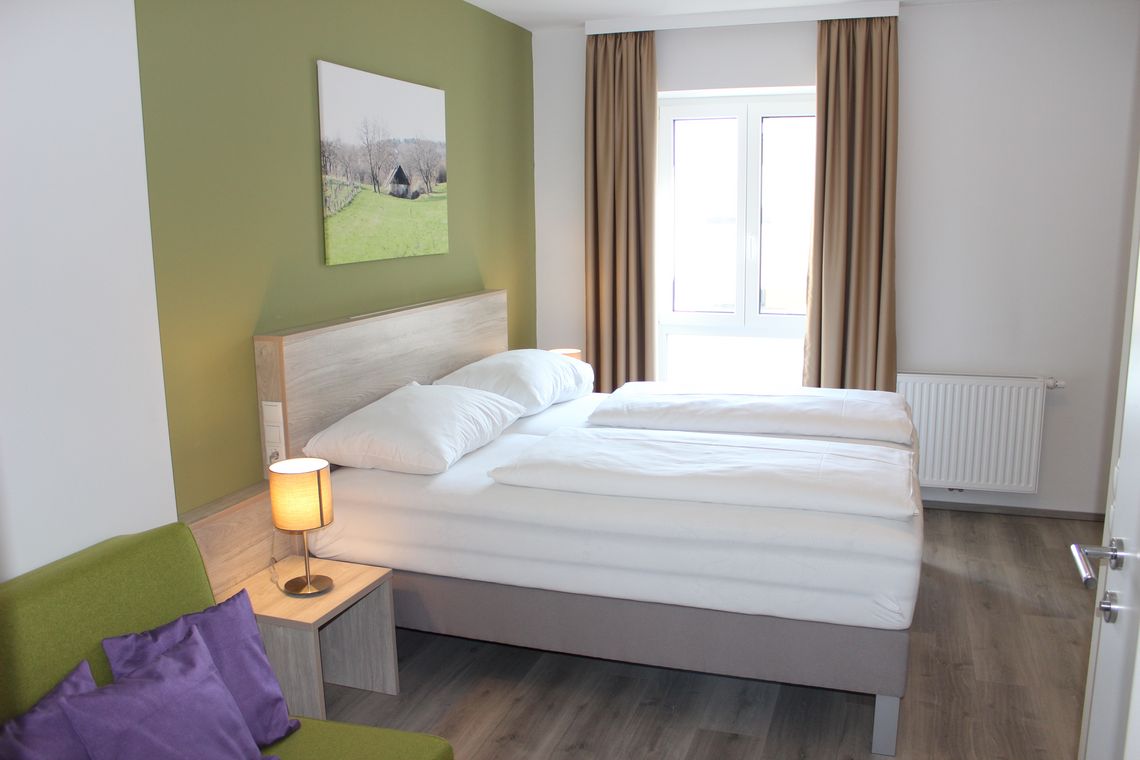 Doppelzimmer mit Doppelbett, grüner Wand und grünem Sessel mit lila Polstern 